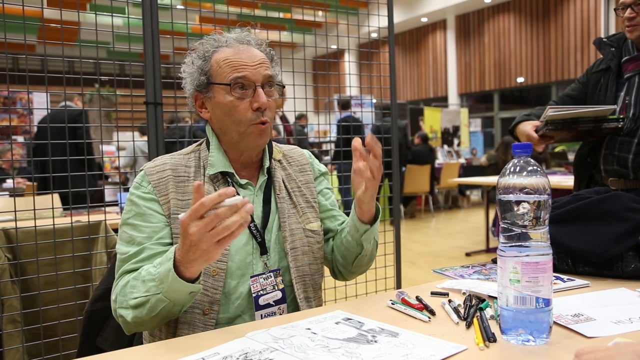 Interview du dessinateur Jean-Pierre Hugo assis à une table et dédicaçant des planches lors d'un festival de la BD.